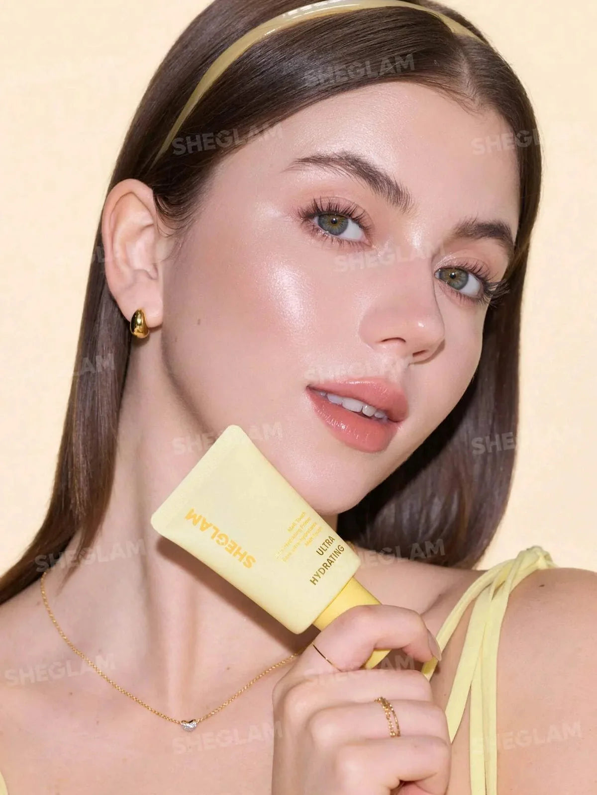 A close-up lifestyle shot of a woman holding a yellow tube of SHEGLAM cream makeup against a warm beige background.
