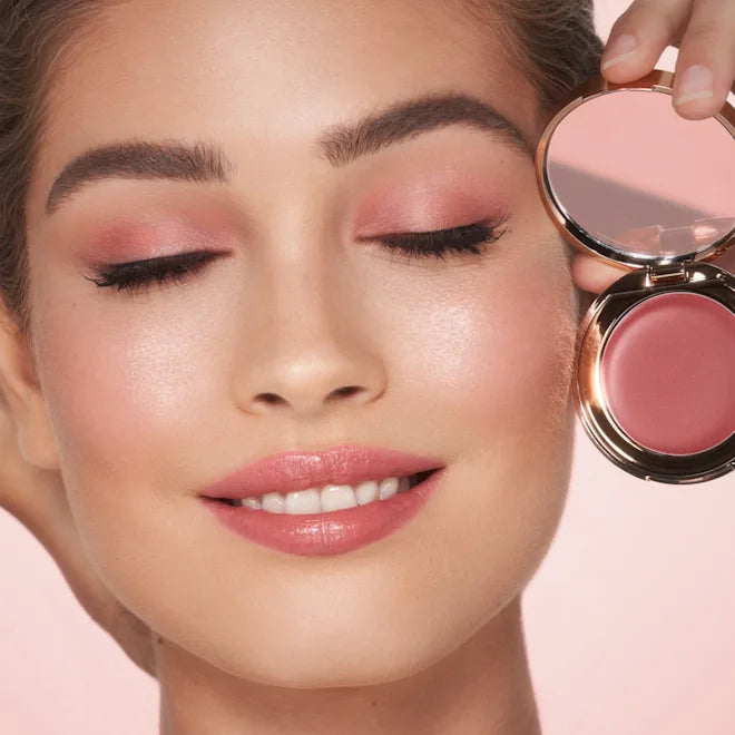 Close-up of a woman applying blush with a compact mirror on a pink background